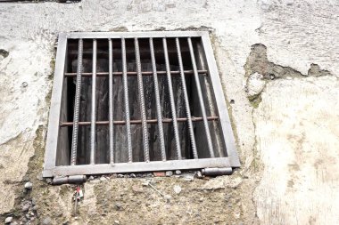 Çürümüş beton kaldırıma metal drenaj ızgarası döşenmiş. Dikdörtgen drenaj kapağı, kenarlarında görünen pas, toprak ve aşınma ile kalın çelik parmaklıklardan yapılmıştır..