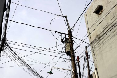 Temiz bir gökyüzüne karşı karmaşık kablolar ve paslı elektrik direkleriyle dolu karmaşık bir küme, kentsel elektrik altyapısının zorluklarını sembolize ediyor. Siyah kabloların yoğun ağı