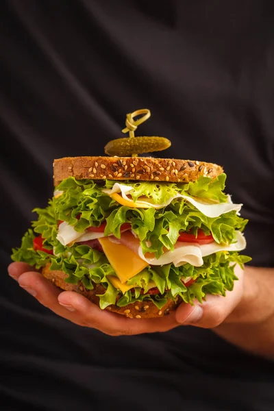 Big sandwich with bread, meat (porshutto), cheese and fresh vegetables in male hands, dark  background.