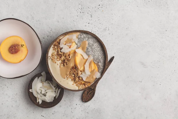 Smoothie bowl with chia pudding, peach, coconut and granola in a coconut bowl on white backgroud, top view. Vegan healthy breakfast, clean eating concept.