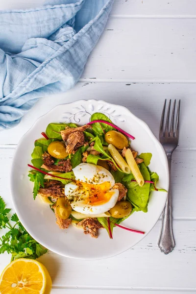 Ton balığı salatası makarna, zeytin ve beyaz ahşap zemin üzerinde beyaz bir tabak içinde haşlanmış yumurta.
