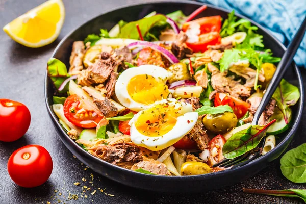 Ton balıklı makarna, zeytin, sebze ve karanlık bir arka plan siyah bir tabağa haşlanmış yumurta salatası.