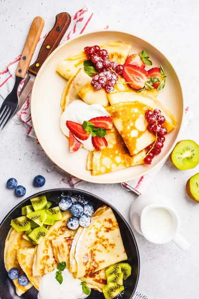 Homemade thin crepes served with curd cream, fruits and berries in black and white plates. Healthy beautiful breakfast concept.