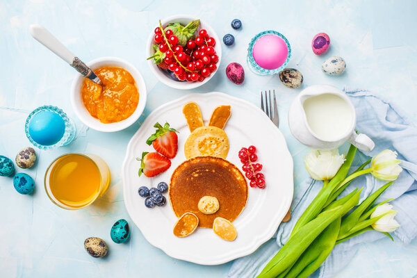 Easter bunny of pancakes with berries. Easter Breakfast table. Colored eggs, milk, juice and jam. Blue background, top view.