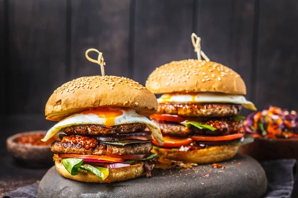 Homemade meat burgers with egg, sauce and vegetables on a dark background.