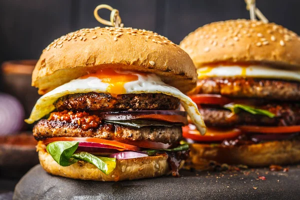 Homemade meat burgers with egg, sauce and vegetables on a dark background.