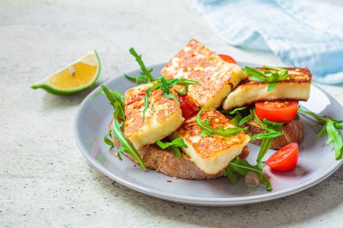 Gri tabakta roka ve domatesli kızarmış halloumi sandviçi. Kızarmış peynir ve domatesli tost.
