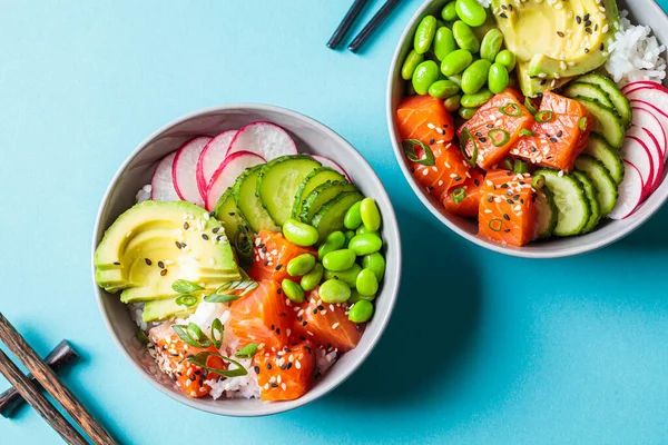 Poke bowl with salmon, rice, avocado, edamame beans, cucumber and radish in a gray bowls, top view. Hawaiian ahi poke bowl, blue background.