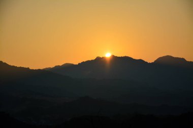 Güneş, Şafak vakti Dağ Sırası 'nda beliriyor: Güneşin, siluetli bir dağ siluetinin üzerinden, ılık tonlu bir sabah gökyüzüne bakan görüntüsü.