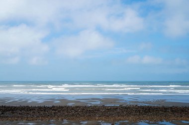 Sörf ve Bulutların Altındaki Çakıl Zeminli Sakin Sahil: Yumuşak dalgaların farklı bulut örtüleri olan bir gökyüzünün altında kıyıda buluştuğu çakıl taşları ve kumsalın huzurlu bir görüntüsü.