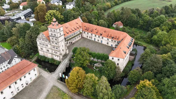 Almanya 'daki Bracke Kalesi' nin belirgin kırmızı çatısı uzaktan çekilen insansız hava aracı görüntüsü. Tarihi kale yemyeşil ağaçlar, çimenli çayırlar ve girişe giden büyüleyici bir köprüsü olan sakin bir nehirle çevrilidir..