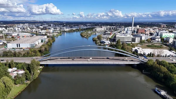 Şehir ve sanayi bölgesinde geniş bir nehri geçen modern bir kemer köprüsünün havadan panoramik görüntüsü. Fotoğrafta şehir binaları, depoları, ofis kompleksleri ve bulutlu açık mavi gökyüzünün altındaki yerleşim bölgeleri görülüyor..