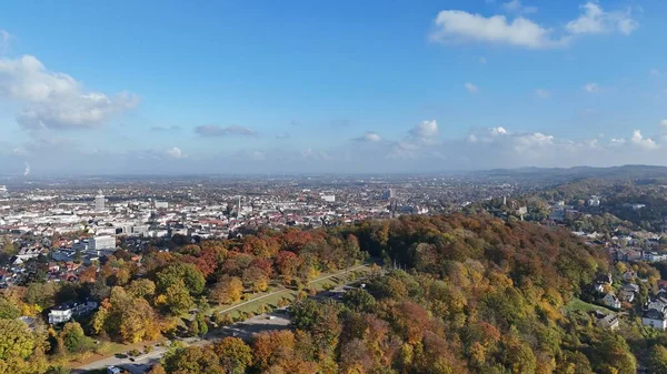 Bielefeld, Almanya 'nın havadan panoramik manzarası, önü renkli sonbahar ormanı ve arkaplanda şehrin ufuk çizgisi. Fotoğraf, şehir mimarisini, tarihi binaları ve açık mavi gökyüzünün altındaki doğal manzarayı gösteriyor.
