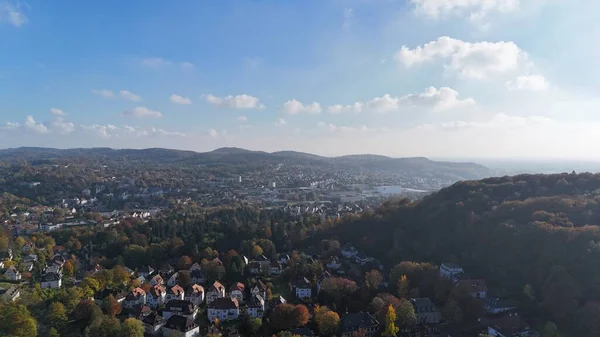 Almanya 'daki Bielefeld' in insansız hava aracı manzarası. Tepeler ve önplanda canlı sonbahar ormanı. Şehrin ufuk çizgisi, yerleşim bölgeleri ve tarihi işaretler dağınık bulutlu parlak bir gökyüzü altında görülebilir.