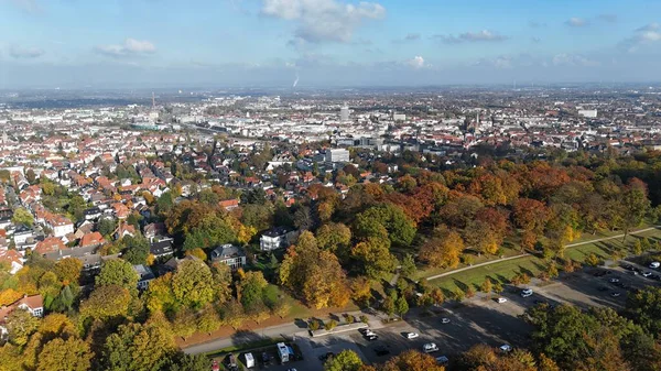 Bielefeld, Almanya 'nın insansız hava aracı görüntüsü, renkli sonbahar ağaçları, büyük bir şehir parkı ve çevresindeki yerleşim yerlerini gösteriyor. Kentsel ufuk çizgisi parlak mavi bir gökyüzünün altında açık bulutlarla görülebilir.