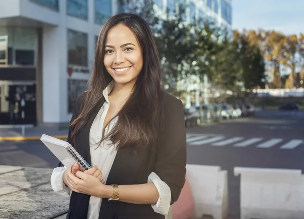 Attractive hispanic young woman in suit teacher, student or secretary standig outdoors with office in the background looking at camera and smiling