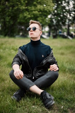 şapkalı adam çimlere oturup güneş gözlüğü takıyor