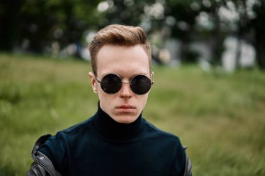 şapkalı adam çimlere oturup güneş gözlüğü takıyor