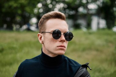 şapkalı adam çimlere oturup güneş gözlüğü takıyor