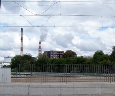 Endüstriyel bacalar bulutlar ve ağaçlarla çevrili kentsel bir ortamda duman yayar. Yakınlardaki binalar, bir şehir manzarasını betimleyen doğal manzarayla tezat oluşturur..