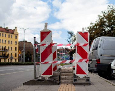 İşçiler kaldırıma inşaat bariyerleri kurarak yayaların geçmesini engelliyor. Yakınlarda gri bir araç park halinde ve bulutlar gökyüzünü dolduruyor..