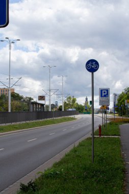 Bir bisiklet şeridi şehir ortamında geniş bir yola paralel gider. Sokak tabelaları, caddelerde ağaçların olduğu park alanlarını gösteriyor. Gökyüzü parçalı bulutlu, sakin bir günü işaret ediyor.
