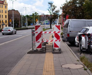 Kaldırıma inşaat bariyerleri kuruldu, yaya girişini kısıtlıyor. Üstü açık mavi gökyüzü olan araçlar caddeye park edilmiş durumda. Bu da şehir ortamında gündüz işi olduğunu gösteriyor..