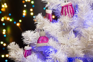 Üzerinde pembe parlak oyuncaklar olan beyaz Noel ağacı. Arka planda parlak bir çelenk var. Bulanık koyu arka plan. Yeni yıl oyuncakları pembe ve gri olarak el ve şapka şeklinde. Yeni Yıl 2020