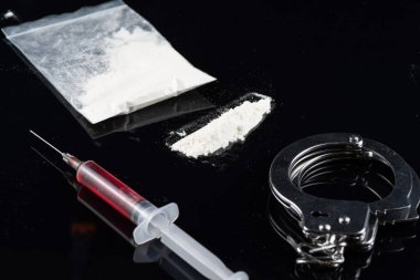 İçinde kırmızı sıvı bulunan bir şırınga, bir plastik beyaz toz torbası, bir çizgi barut, ve karanlık bir yüzeye kelepçeler..