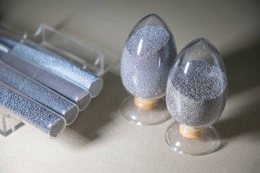 Dört uzun cam tüp ve iki farklı yumurta şeklindeki cam kavanozda bir dizi çelik mermi veya tanecikli malzeme sergileniyor..