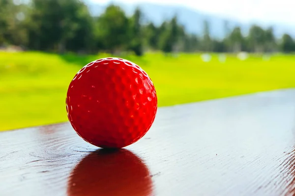 Golf red ball on a wooden surface, green field and blue sky. Macro view, luxury sport concept.
