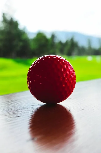 Golf red ball on a wooden surface, green field and blue sky. Macro view, luxury sport concept.