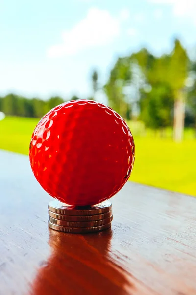 Golf red ball on a pile of coins, green grass field and blue sky background. Macro view, luxury sport game concept.