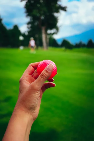 Golf pink ball hold in a woman's hand, green course grass, blue sky.
