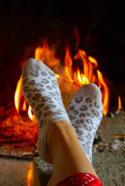 Burning wood at the fireplace, female legs in socks warming up. Firewood bricks at the fire, woman foot heating. clipart