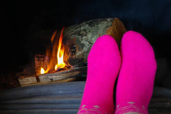 Burning wood at the fireplace, female feet in pink socks warming up. Firewood bricks at the fire, woman foot heating.