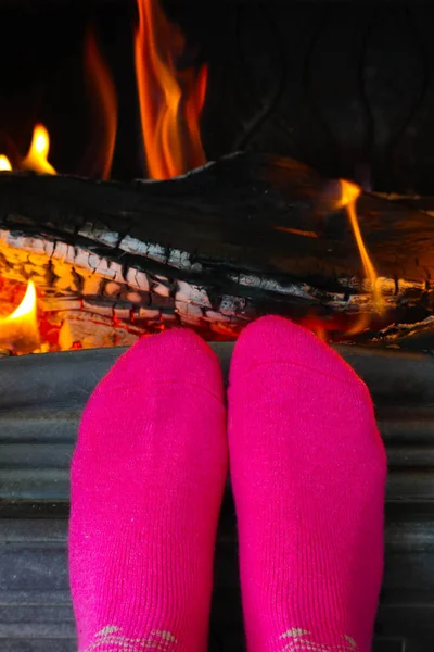 Burning wood at the fireplace, female feet in pink socks warming up. Firewood bricks at the fire, woman foot heating.