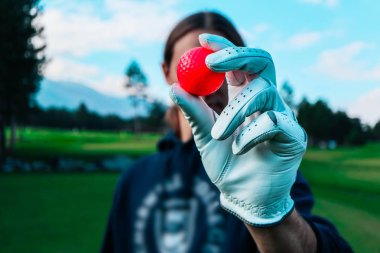 Bir el bir kırmızı golf topu gösterilen genç golfçü beyaz eldiven ile kapatın. Yeşil sahaları, mavi gökyüzü ve arka planda dağlar.