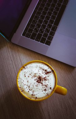 Cappuccino kahve ofis ahşap masa üzerinde laptop klavye arka planda büyük bir sarı bardak.