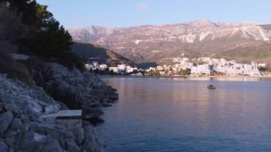 Denizin üzerindeki kayalıklar ve berrak mavi gökyüzünün altında, dağların eteklerindeki beyaz, güzel tatil köyünün büyüleyici panoramik manzarası. Akdeniz turistik bölgesi, Budva, Karadağ.