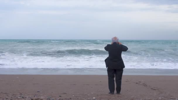 Літній сірошерстий чоловік у костюмі стоїть на самоті на березі штормового моря в погану вітряну погоду, фотографує пейзаж. Депресія і самотність пенсіонерів — стокове відео