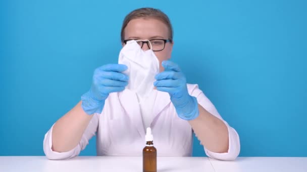 Giovane donna con camice medico bianco e guanti protettivi blu seduta al tavolo di lavoro con bottiglia di vetro scuro, prende la maschera facciale e indossarla. Laboratorio o medico al lavoro. — Video Stock
