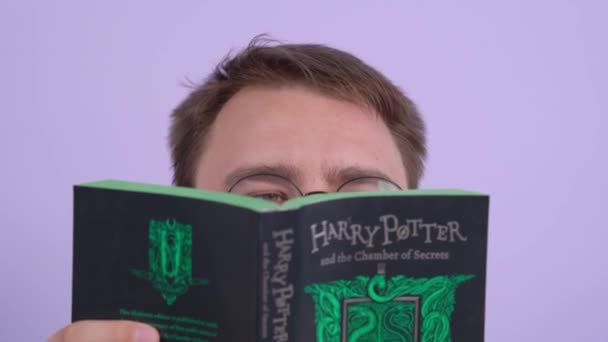 Rostov, Rusko 20. července 2020: Portrét mladého muže, který čte román Harryho Pottera a Tajemné komnaty a v obou rukou drží knihu. Na něco se podívá přes brýle a zavrtí hlavou. — Stock video