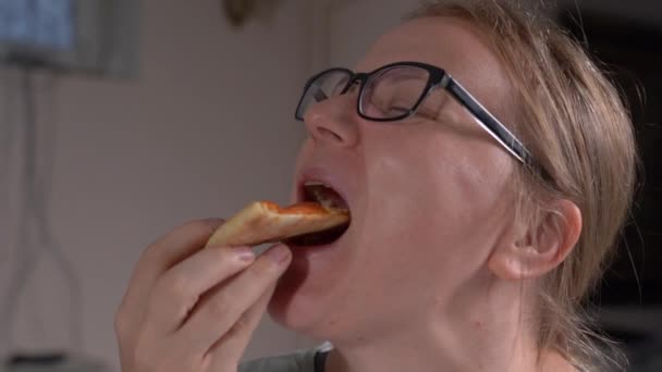 Gözlüklü genç sarışın kadın bir dilim baharatlı pepperoni pizzasını ısırıyor ve zevkle çiğniyor, yaklaşın, yavaş çekim. Sağlıksız yaşam tarzı ve oburluk — Stok video