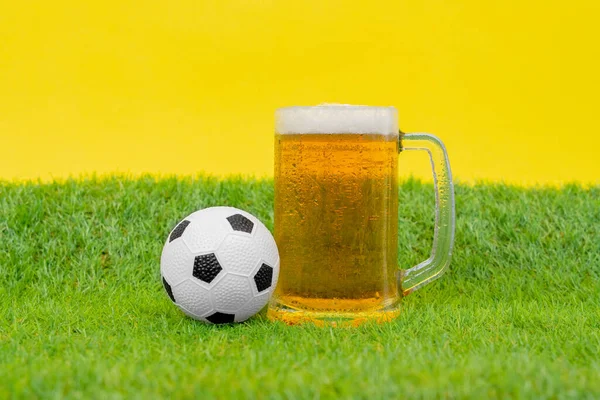 Taza grande llena de cerveza fría con espuma, y soporte de pelota de fútbol en miniatura sobre hierba de césped artificial sobre fondo amarillo. Atributos del pasatiempo y entretenimiento masculino tradicional — Foto de Stock
