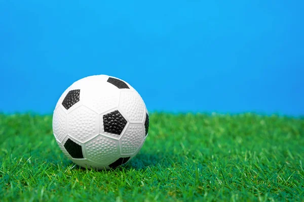 Pelota de fútbol de juguete en miniatura se encuentra en la hierba verde de césped artificial del campo de fútbol, fondo azul, vista frontal, de cerca. Equipamiento para actividades al aire libre y deportes — Foto de Stock