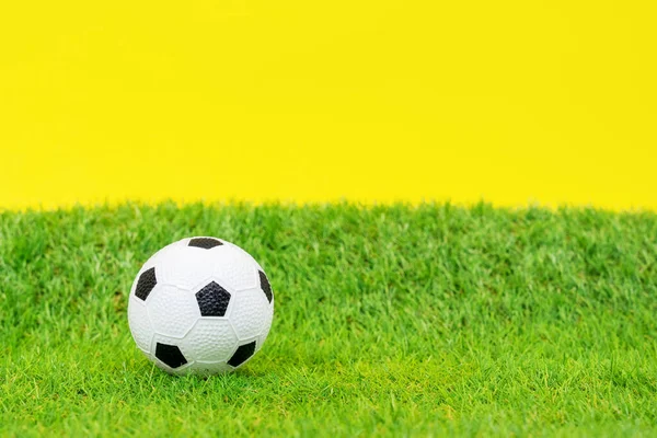 Pelota de fútbol de juguete en miniatura se encuentra en la hierba verde de césped artificial del campo de fútbol, fondo amarillo, vista frontal, de cerca. Equipamiento para actividades al aire libre y deportes — Foto de Stock