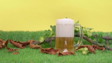 Büyük bardak bira, kalın köpük ve sarı arka planla dolu. Kupa, kuru yapraklar, dallar ve atlama kümeleriyle süslenmiş yapay çimenlerin üzerinde duruyor.
