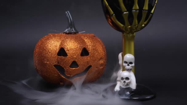 Mysterious smoke creeps over the surface on which there are attributes of Halloween party - shiny jack lantern pumpkin and goblet decorated with skulls and skeleton hand, front view — Stock Video