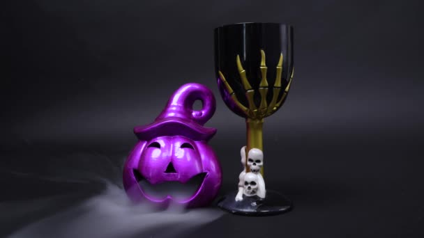 Calabaza linterna jack púrpura con sonrisa siniestra y copa espeluznante con cráneos y mano esqueleto para decorar la habitación para la fiesta de Halloween sobre fondo negro, nubes de humo misterioso alrededor — Vídeos de Stock